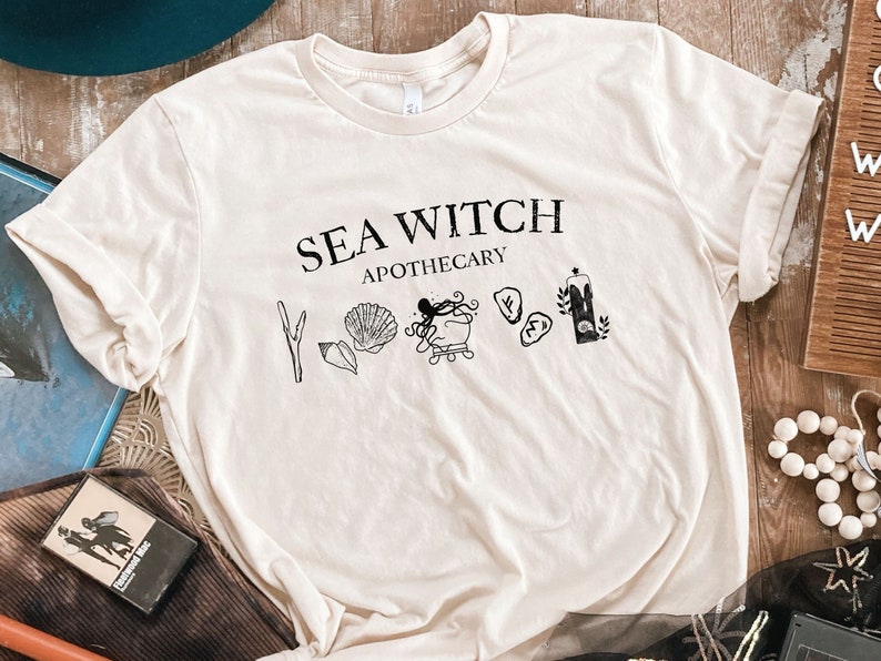 May include: A beige t-shirt with the text "SEA WITCH APOTHECARY" printed in black. The design includes a variety of nautical symbols, including an octopus, seashells, and a candle.