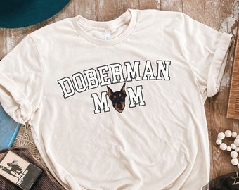 May include: A cream-colored t-shirt with the text "Doberman Mom" in black outlined letters. A black and brown Doberman Pinscher head is centered below the text.