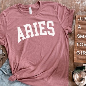 K&ouml;nnte beinhalten: Ein staubiges rosafarbenes T-Shirt mit dem Wort "ARIES" in wei&szlig; auf der Vorderseite. Das Shirt ist gefaltet und auf einer Holzoberfl&auml;che abgelegt.
