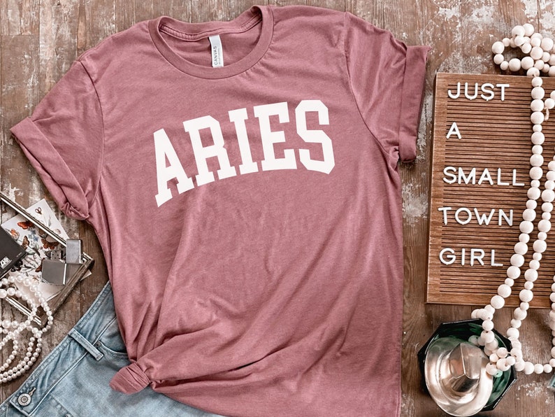K&ouml;nnte beinhalten: Ein staubiges rosafarbenes T-Shirt mit dem Wort "ARIES" in wei&szlig; auf der Vorderseite. Das Shirt ist gefaltet und auf einer Holzoberfl&auml;che abgelegt.