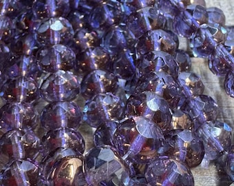 May include: Close-up of a collection of faceted, round beads in shades of purple and iridescent hues. The beads have a reflective, glass-like appearance, suggesting they are made of crystal or a similar material. The beads are strung together.