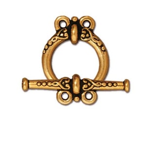 May include: Gold-tone metal toggle clasp with an ornate design. The clasp has a round ring and a bar with a decorative pattern.