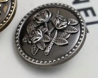 May include: A silver-toned metal button with a raised design of three roses and leaves. The button has a beaded edge.