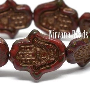 May include: Close-up of several red-brown Hamsa hand beads with a detailed design. The beads have a metallic finish and are strung together. The text "Nirvana Beads" is visible in the image.