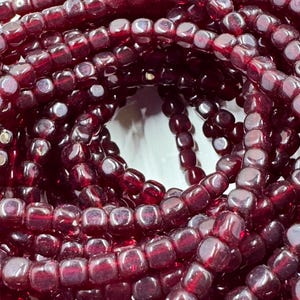 May include: Close-up of multiple strands of dark red, square-shaped beads. The beads are strung together, creating a tangled, layered effect. The beads have a glossy finish, reflecting light and adding depth to the image. Ideal for jewelry making.