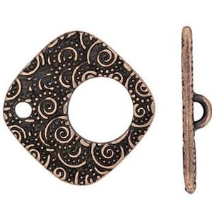 May include: Bronze-toned toggle clasp with an ornate, swirling pattern. The square-shaped clasp features a central circular opening and a smaller hole. The matching bar has a loop on one end. The clasp is approximately 1 inch wide.