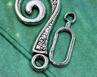 May include: Silver-toned metal clasp with an ornate, swirling design. The clasp features a decorative hook and a rectangular loop, perfect for jewelry making or crafting projects. The clasp is set against a green background.