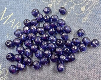 May include: A collection of faceted, dark blue beads. The beads are scattered across a light blue fabric with the word "MAR" visible. The beads have a glossy finish and are round with multiple facets.