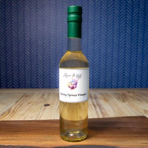Peut inclure: Une bouteille en verre transparent de vinaigre d'épinette de printemps avec un bouchon vert. L'étiquette porte le texte "Mama Witch" et une illustration à l'aquarelle. La bouteille est posée sur une surface en bois devant un fond bleu à motifs.