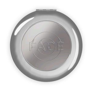 Puede incluir: Un espejo compacto plateado con la palabra "FACE" grabada en la parte delantera. El compacto es redondo con una tapa con bisagras y un acabado metálico brillante. El diseño presenta círculos concéntricos.