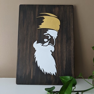 May include: A wooden wall art piece featuring a white silhouette of a person with a beard and a gold turban. The background is a dark brown wood.