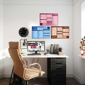 May include: A home office workspace featuring a black desk, a brown leather office chair, a computer monitor, a black clock, and three posters on the wall. The posters are blue, pink, and brown and display text and graphics related to Adobe Photoshop, Illustrator, and InDesign.