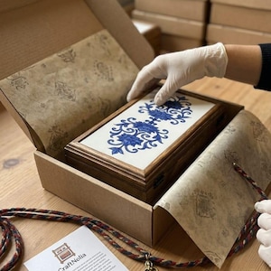 May include: A rectangular wooden box with a white lid featuring a blue floral pattern, nestled inside an open cardboard shipping box lined with patterned tissue paper. A white card with the text CraftNolia and thank you is placed nearby.
