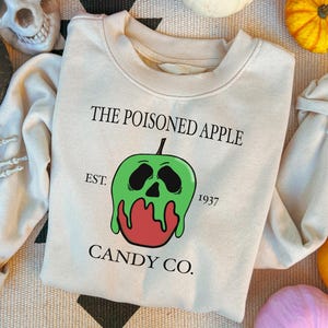 May include: A cream-coloured sweatshirt featuring the text "THE POISONED APPLE CANDY CO." and a graphic of a green apple with a skull design. The sweatshirt also has the text "EST. 1937". Halloween decorations surround the sweatshirt.