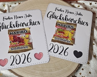 Può includere: Due cartoncini rettangolari bianchi con il testo Frohes Neues Jahr e Glücksbärchen, ciascuno fissato con un sacchetto di caramelle gommose Haribo Goldbären. Le carte riportano l'anno 2026 in nero, con due cuori rosa o un cuore nero.