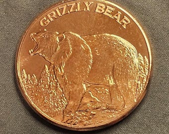 May include: A round copper-colored coin featuring a raised relief of a roaring grizzly bear standing in a forest landscape. The text GRIZZLY BEAR is embossed in a curved arc along the top edge of the coin, which has a metallic finish.