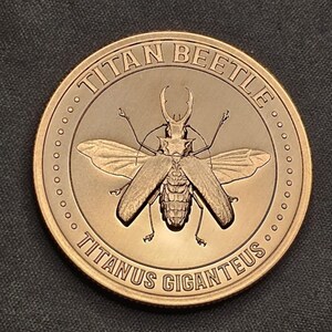 May include: A bronze-colored coin featuring a detailed depiction of a Titan Beetle with outstretched wings. The coin's edge is adorned with small circles, and the text "TITAN BEETLE" and "TITANUS GIGANTEUS" are inscribed around the beetle.