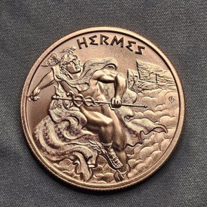 May include: A bronze-colored coin featuring the word "HERMES" at the top. The coin depicts a muscular figure holding a staff, with wings on his head and feet. The background includes clouds and a flag.