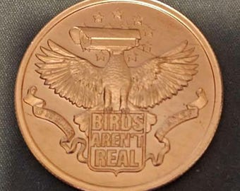 May include: A copper-colored coin with an eagle design, wings spread, and a surveillance camera head. The text "BIRDS AREN'T REAL" is centered below the eagle. The coin has a decorative border and stars at the top.