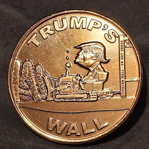 May include: A bronze-colored coin with the words "TRUMP'S WALL" in a circular design. The coin features a caricature of a man operating a bulldozer, with the text "BORDER WALL - DO NOT CROSS".