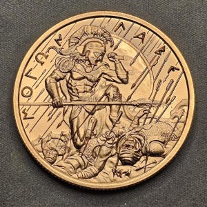 May include: A bronze-colored coin featuring a detailed relief of a warrior in a helmet, wielding a spear and sword, with fallen enemies. The coin has Greek lettering around the edge and depicts a battle scene.