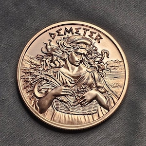 May include: A circular bronze-toned commemorative coin featuring a relief of Demeter holding a basket of wheat and fruit. The figure has flowing hair and is set against a landscape background. The text DEMETER is inscribed in a stylized font at the top.