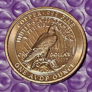 May include: A gold-colored coin with an eagle design. The coin has text that reads "COPPER .999 FINE", "UNITED STATES OF AMERICA", "ONE DOLLAR", and "ONE AVDP OUNCE". The coin is on a purple background.
