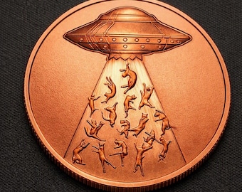 May include: A circular copper-toned coin featuring an embossed design of a flying saucer emitting a beam of light. Inside the light beam, multiple cats are depicted in various poses as if being abducted or floating upward toward the spacecraft.
