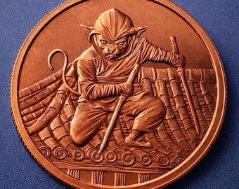 May include: A circular copper-toned coin featuring a relief design of a cat dressed in a hooded ninja outfit. The cat is crouched on a tiled roof, holding a long sword in one hand while its tail curls upward. The coin has a serrated edge.