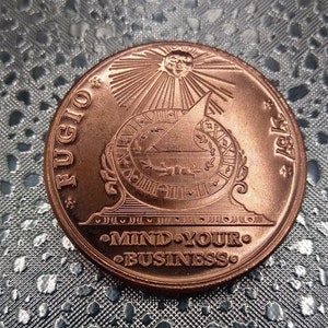 May include: A round copper-colored coin featuring a sundial design with a sun above it. The text FUGIO and 1787 are inscribed on the sides, while the words MIND YOUR BUSINESS are embossed at the bottom of the coin face.