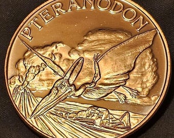 May include: A bronze-colored coin featuring a detailed relief of a Pteranodon in flight over a prehistoric landscape. The word "PTERANODON" is inscribed along the top edge. The coin's design includes a volcano and ancient structures.