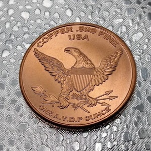 Puede incluir: Una moneda redonda de cobre con un dise&ntilde;o de &aacute;guila. La moneda dice "Copper .999 Fine USA One A.V.D.P Ounce".