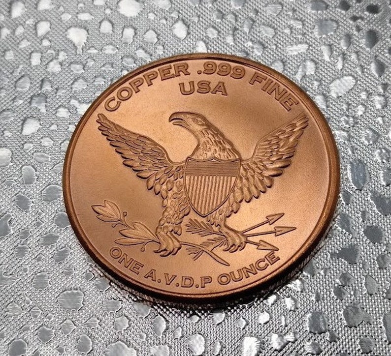 Puede incluir: Una moneda redonda de cobre con un dise&ntilde;o de &aacute;guila. La moneda dice "Copper .999 Fine USA One A.V.D.P Ounce".