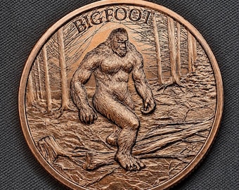 May include: A round copper-colored coin featuring a detailed relief of a hairy Bigfoot creature walking through a forest. The word BIGFOOT is embossed in an arc at the top of the coin. The design shows trees, a fallen log, and textured ground.