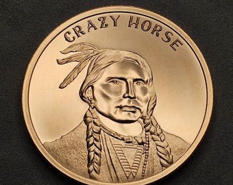 May include: A bronze-colored coin featuring a detailed relief of Crazy Horse, a Native American war leader. The coin's edge has the words "CRAZY HORSE" in an arch. The portrait shows Crazy Horse with braided hair and a feathered headdress.