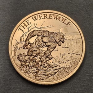 May include: A bronze-colored coin featuring a detailed relief of a werewolf in a menacing pose. The coin's edge has the words "THE WEREWOLF" and a background with a full moon, trees, and a town.