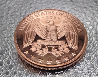 May include: A copper-colored coin featuring an eagle with outstretched wings, surrounded by the words "UNITED STATES OF AMERICA." The coin has a detailed design with stars and laurel branches. The coin is round and has a textured edge.