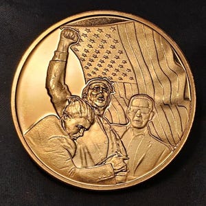 May include: A bronze-colored commemorative coin featuring a relief of three figures with an American flag. One figure raises a fist, another holds the flag, and a third figure is in the foreground. The coin has a circular shape.