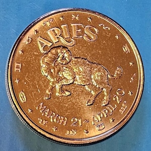 May include: A round copper-toned medallion featuring a central embossed ram figure. The word ARIES is arched at the top, with the text MARCH 21 - APRIL 20 at the bottom. The outer rim is decorated with various small zodiac symbols and stars.