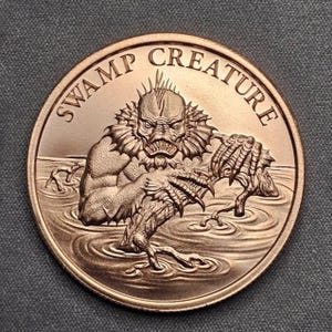 May include: Bronze-colored coin featuring a detailed relief of a swamp creature emerging from water. The creature has a menacing expression and clawed hands. The words "SWAMP CREATURE" are inscribed around the coin's edge.