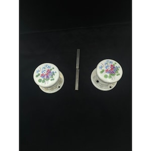 May include: Two white ceramic door knobs with floral designs in blue, purple, and pink. The knobs have a gold trim and are accompanied by a metal latch. The knobs are likely for a cabinet or drawer.