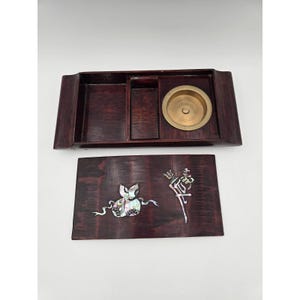 May include: A rectangular dark wood box with a removable lid featuring iridescent mother-of-pearl inlay of a gourd and calligraphy. The interior contains two open compartments and a circular brass insert, providing a functional storage space.