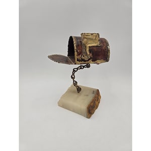 May include: A handcrafted mailbox sculpture with a hinged lid, made of copper and bronze. The mailbox is mounted on a chain and a rectangular stone base. The sculpture has a rustic, vintage aesthetic.