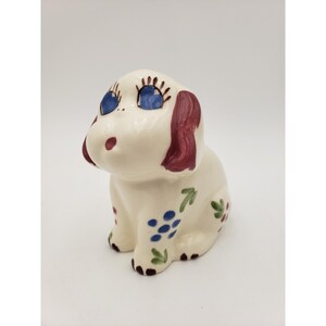 May include: A ceramic dog figurine in a sitting position. The dog is primarily white with blue eyes, red ears, and a red nose. It has blue and green floral accents. The figurine has a whimsical, cartoonish design.