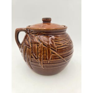 May include: A brown ceramic pot with a lid and handle. The pot has a round shape with a textured surface and a decorative leaf pattern. The lid has a small knob. The pot is likely used for cooking or storage.