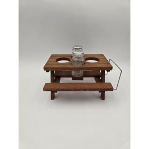 May include: A miniature wooden picnic table condiment holder with a dark brown finish. The table features two circular cutouts on the tabletop, one of which holds a clear glass salt or pepper shaker with a silver metal screw-on lid.
