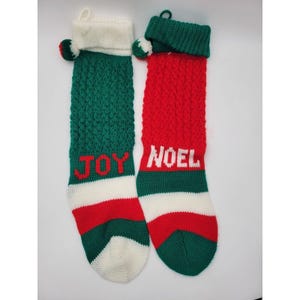 May include: Two knitted Christmas stockings. One is green with the word "JOY" in red, and the other is red with the word "NOEL" in white. Both have white cuffs and bottoms, and green and red pom-poms.