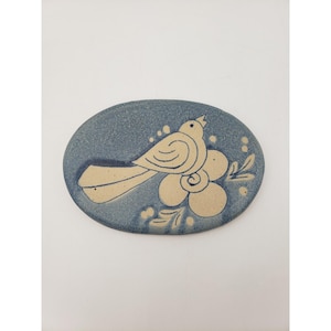 May include: Oval ceramic soap dish with a blue background and a beige bird and flower design. The bird is perched on a flower with leaves. The dish has a handmade, artisanal feel.
