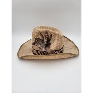 May include: A straw cowboy hat with a wide brim. The hat is tan with a black and brown striped band and decorative feathers. The hat is a neutral color and suitable for various outfits.