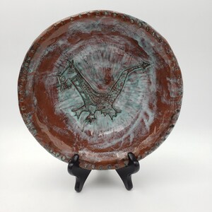 May include: A handmade ceramic plate with a dragon design. The plate features a brown exterior with a turquoise dragon in the center. The rim has a dotted pattern. The plate is displayed on a black stand.